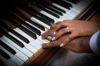 Two hands gently rest on a piano keyboard, one adorned with an elegant engagement ring and the other wearing a wedding band. The piano keys are black and white, suggesting a classic, timeless setting.
