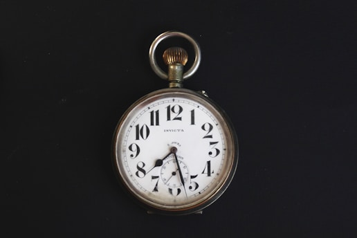 A vintage pocket watch with a white face, black numerals, and a silver casing. The watch has ornate hands and a loop at the top for attaching a chain. It is set against a stark black background, highlighting its classic design.