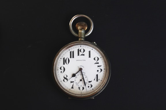 A vintage pocket watch with a white face, black numerals, and a silver casing. The watch has ornate hands and a loop at the top for attaching a chain. It is set against a stark black background, highlighting its classic design.