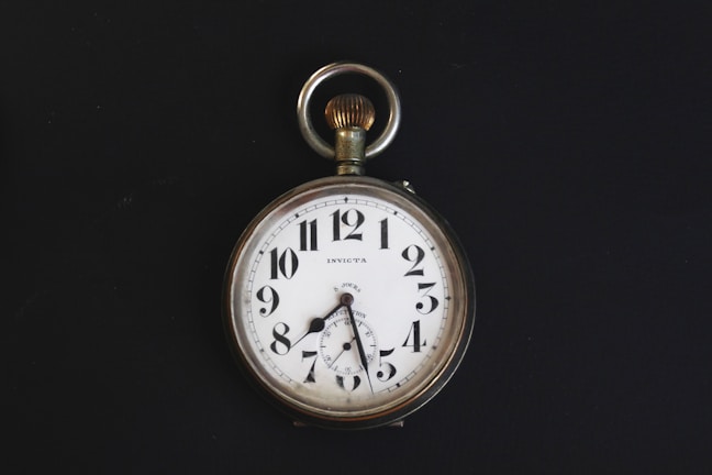 A vintage pocket watch with a white face, black numerals, and a silver casing. The watch has ornate hands and a loop at the top for attaching a chain. It is set against a stark black background, highlighting its classic design.