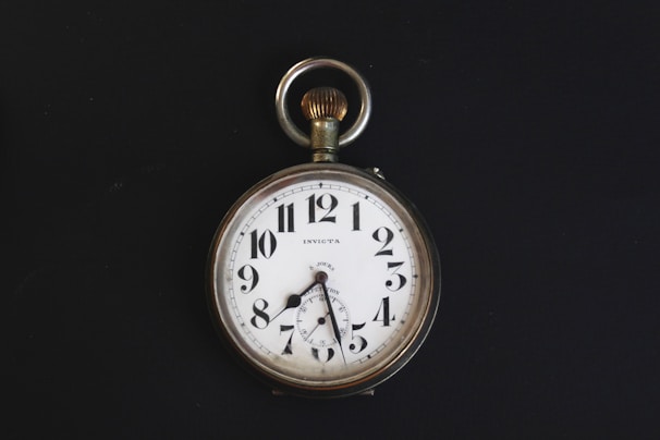 A vintage pocket watch with a white face, black numerals, and a silver casing. The watch has ornate hands and a loop at the top for attaching a chain. It is set against a stark black background, highlighting its classic design.