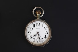 A vintage pocket watch with a white face, black numerals, and a silver casing. The watch has ornate hands and a loop at the top for attaching a chain. It is set against a stark black background, highlighting its classic design.