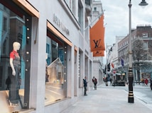 A high-end shopping street with a prominent Louis Vuitton store. Large windows display mannequins dressed in stylish clothing. The street is lined with upscale boutiques, flags, and street lamps. A few pedestrians can be seen walking or standing, some checking their phones.