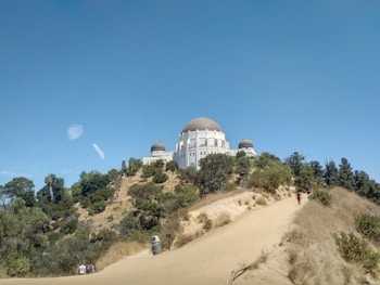 A white observatory building with a large dome is situated on a hill surrounded by greenery and a clear blue sky. A path leads up the hill with a few people walking, enjoying a sunny day.