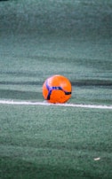 A vibrant orange and blue soccer ball rests near a white line on a green synthetic turf field. The scene is well-lit, highlighting the texture of the turf and the colors of the ball.