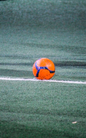 A vibrant orange and blue soccer ball rests near a white line on a green synthetic turf field. The scene is well-lit, highlighting the texture of the turf and the colors of the ball.