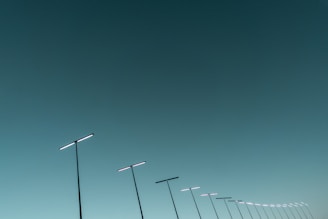 A series of sleek, modern streetlights are aligned diagonally against a vast, clear sky. The streetlights have a minimalist design, featuring long poles with horizontal light bars.