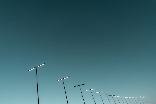 A series of sleek, modern streetlights are aligned diagonally against a vast, clear sky. The streetlights have a minimalist design, featuring long poles with horizontal light bars.