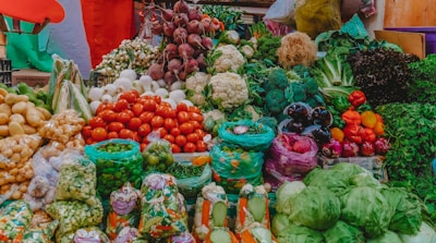 A vibrant display of various fresh vegetables at a market. The assortment includes tomatoes, cauliflower, beetroots, potatoes, onions, green beans, capsicums, cabbages, and other leafy greens. The produce is arranged in an aesthetically pleasing, colorful manner, with some vegetables packed in plastic bags while others are arranged in open piles. The background includes some multi-colored fabric that adds to the lively ambiance of the marketplace.