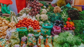 A vibrant display of various fresh vegetables at a market. The assortment includes tomatoes, cauliflower, beetroots, potatoes, onions, green beans, capsicums, cabbages, and other leafy greens. The produce is arranged in an aesthetically pleasing, colorful manner, with some vegetables packed in plastic bags while others are arranged in open piles. The background includes some multi-colored fabric that adds to the lively ambiance of the marketplace.