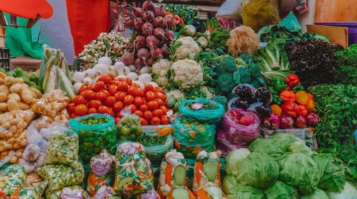 A vibrant display of various fresh vegetables at a market. The assortment includes tomatoes, cauliflower, beetroots, potatoes, onions, green beans, capsicums, cabbages, and other leafy greens. The produce is arranged in an aesthetically pleasing, colorful manner, with some vegetables packed in plastic bags while others are arranged in open piles. The background includes some multi-colored fabric that adds to the lively ambiance of the marketplace.