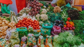 A vibrant display of various fresh vegetables at a market. The assortment includes tomatoes, cauliflower, beetroots, potatoes, onions, green beans, capsicums, cabbages, and other leafy greens. The produce is arranged in an aesthetically pleasing, colorful manner, with some vegetables packed in plastic bags while others are arranged in open piles. The background includes some multi-colored fabric that adds to the lively ambiance of the marketplace.