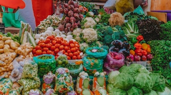 A vibrant display of various fresh vegetables at a market. The assortment includes tomatoes, cauliflower, beetroots, potatoes, onions, green beans, capsicums, cabbages, and other leafy greens. The produce is arranged in an aesthetically pleasing, colorful manner, with some vegetables packed in plastic bags while others are arranged in open piles. The background includes some multi-colored fabric that adds to the lively ambiance of the marketplace.