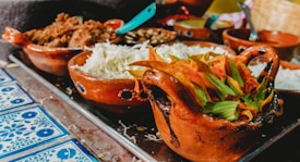 A variety of traditional Mexican dishes on a tray, placed on a decorative tiled table. Clay bowls are filled with ingredients such as shredded cabbage and colorful squash blossoms, accompanied by other items like seasoned meat and sauces. The vivid colors and textures contribute to an inviting and vibrant presentation.