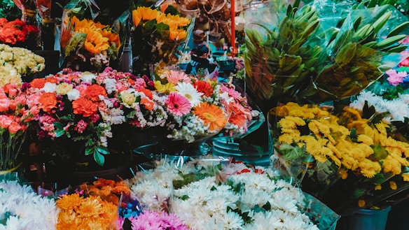 A vibrant display of various flowers, including sunflowers, daisies, carnations, and chrysanthemums, organized in bouquets. Many of the flowers are wrapped in clear plastic and arranged in a crowded, colorful assortment. The scene suggests a bustling flower market or shop, with a focus on the richness and variety of the floral arrangements.