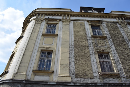 The image features an old building with an ornate facade. The exterior walls are aged, with patches of ivy clinging to the surface, giving it a historic appearance. Large windows with intricate frames dominate the structure, and a few sections show signs of wear and peeling paint. The sky is clear with a few clouds visible, adding contrast to the building's pale tones.