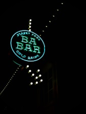 A neon sign reading 'Street Food BA BAR Cold Drink' illuminated against a dark background. The sign is round, with 'BA BAR' prominently displayed in the center, and small lightbulbs outlining the structure. The neon glow gives off a vibrant, inviting aura.