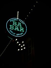 A neon sign reading 'Street Food BA BAR Cold Drink' illuminated against a dark background. The sign is round, with 'BA BAR' prominently displayed in the center, and small lightbulbs outlining the structure. The neon glow gives off a vibrant, inviting aura.