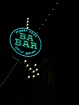 A neon sign reading 'Street Food BA BAR Cold Drink' illuminated against a dark background. The sign is round, with 'BA BAR' prominently displayed in the center, and small lightbulbs outlining the structure. The neon glow gives off a vibrant, inviting aura.