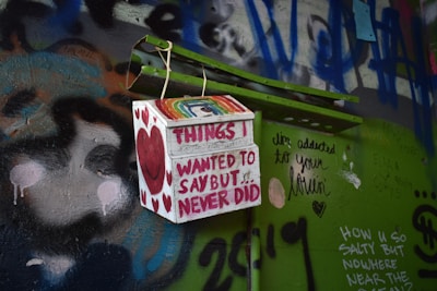 A colorful and graffiti-covered wall features a small box with the words 'Things I wanted to say but never did' written on it. The box displays a smiling heart and rainbow design. Surrounding the box are various graffiti texts and drawings, including a message about addiction to love and other personal notes from different people.
