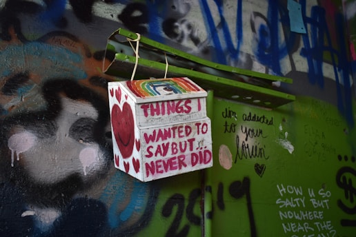 A colorful and graffiti-covered wall features a small box with the words 'Things I wanted to say but never did' written on it. The box displays a smiling heart and rainbow design. Surrounding the box are various graffiti texts and drawings, including a message about addiction to love and other personal notes from different people.