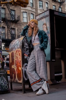 A person leans casually against a graffiti-covered mailbox on a city street, wearing a yellow beanie, teal jacket, white crop top, plaid wide-leg pants, and white sneakers. The backdrop includes an older building with fire escapes, and a postered phone booth.
