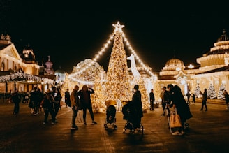 A festive scene with a large Christmas tree adorned with bright, twinkling lights. People are gathered around, some with strollers, enjoying the illuminated surroundings. The architecture in the background is also decorated with lights, adding to the cheerful atmosphere.