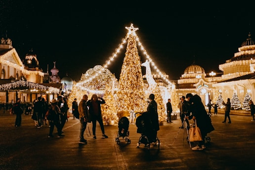 A festive scene with a large Christmas tree adorned with bright, twinkling lights. People are gathered around, some with strollers, enjoying the illuminated surroundings. The architecture in the background is also decorated with lights, adding to the cheerful atmosphere.