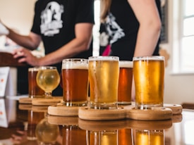 Five glasses of different types of beer are arranged on wooden coasters on a reflective table. Two people in the background, partially out of focus, are dressed in casual attire.