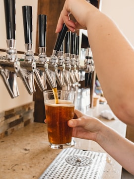 A hand is pulling a tap handle on a row of beer taps, filling a glass with amber-colored beer. The setting appears to be a bar or a brewery with a countertop in the foreground.