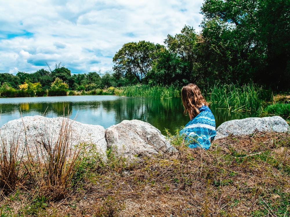 woman in blue and white dress sitting on rock near lake during daytime