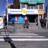A street corner with a pharmacy named 'Farmacias Bolivia' on a sunny day. The building has colorful signage, and a few people are seen walking and sitting near the entrance. A crosswalk is visible in the foreground, and the scene includes several small posters and advertisements around the entrance.