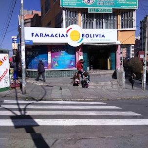 A street corner with a pharmacy named 'Farmacias Bolivia' on a sunny day. The building has colorful signage, and a few people are seen walking and sitting near the entrance. A crosswalk is visible in the foreground, and the scene includes several small posters and advertisements around the entrance.