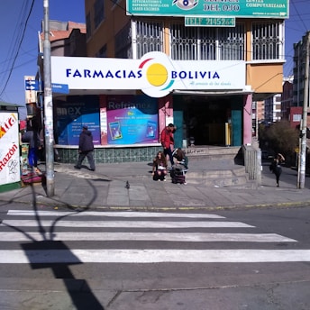 A street corner with a pharmacy named 'Farmacias Bolivia' on a sunny day. The building has colorful signage, and a few people are seen walking and sitting near the entrance. A crosswalk is visible in the foreground, and the scene includes several small posters and advertisements around the entrance.