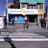 A street corner with a pharmacy named 'Farmacias Bolivia' on a sunny day. The building has colorful signage, and a few people are seen walking and sitting near the entrance. A crosswalk is visible in the foreground, and the scene includes several small posters and advertisements around the entrance.