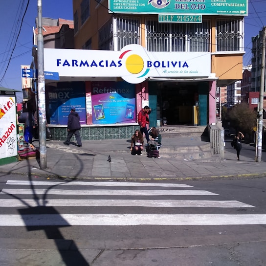 A street corner with a pharmacy named 'Farmacias Bolivia' on a sunny day. The building has colorful signage, and a few people are seen walking and sitting near the entrance. A crosswalk is visible in the foreground, and the scene includes several small posters and advertisements around the entrance.