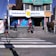 A street corner with a pharmacy named 'Farmacias Bolivia' on a sunny day. The building has colorful signage, and a few people are seen walking and sitting near the entrance. A crosswalk is visible in the foreground, and the scene includes several small posters and advertisements around the entrance.