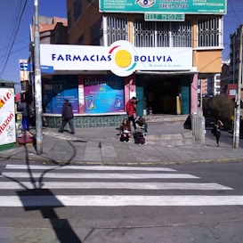 A street corner with a pharmacy named 'Farmacias Bolivia' on a sunny day. The building has colorful signage, and a few people are seen walking and sitting near the entrance. A crosswalk is visible in the foreground, and the scene includes several small posters and advertisements around the entrance.