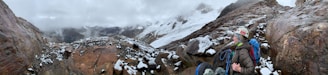 A hiker wearing climbing gear and a helmet stands on rocky terrain covered in patches of snow. The background features a rugged mountain landscape with snow-covered peaks and a cloudy sky.