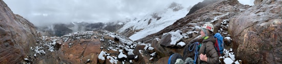 A hiker wearing climbing gear and a helmet stands on rocky terrain covered in patches of snow. The background features a rugged mountain landscape with snow-covered peaks and a cloudy sky.