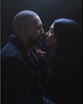 A couple is engaged in an intimate moment, with their faces close and a hand gently touching the man's chin. The setting is dimly lit, creating a dramatic and tender atmosphere.
