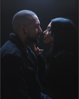 A couple is engaged in an intimate moment, with their faces close and a hand gently touching the man's chin. The setting is dimly lit, creating a dramatic and tender atmosphere.