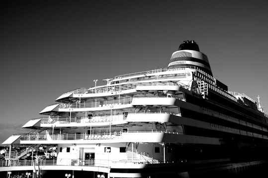 A large cruise ship with multiple decks and a sleek, modern design is captured against a clear sky. The ship's structure includes several balconies and railings, showcasing its expansive and luxurious construction.
