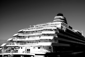 A large cruise ship with multiple decks and a sleek, modern design is captured against a clear sky. The ship's structure includes several balconies and railings, showcasing its expansive and luxurious construction.
