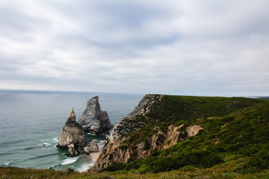 green and black rock formation on body of water under white clouds during daytime in Sintra-Cascais Natural Park Portugal