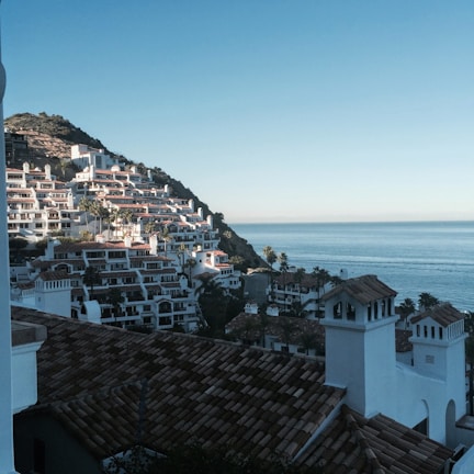 A picturesque coastal hillside is densely populated with white, Mediterranean-style buildings featuring terracotta roofs. Palm trees are sprinkled throughout the area, adding to the scenic beauty. The setting sun casts a warm glow, and the expansive blue ocean stretches into the horizon.