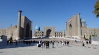 A large, open square surrounded by grand, ornately decorated buildings with tall arches and turquoise domes under a clear blue sky. Many people are walking or standing in the square, some in groups and others taking photos. The architecture is rich with intricate patterns and designs.