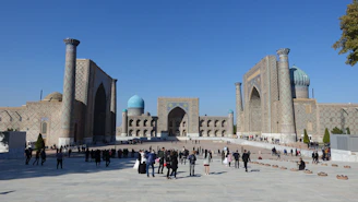 A professional local guide warmly engaging with travelers in front of the majestic Registan Square under a clear blue sky.
