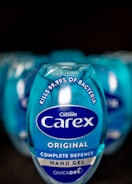A close-up view of a blue Carex hand gel bottle prominently displaying text that states it kills 99.99% of bacteria. The bottle design includes the Carex logo and branding elements on a shiny, reflective surface. Multiple bottles are blurred in the background, suggesting a commercial arrangement.