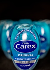 A close-up view of a blue Carex hand gel bottle prominently displaying text that states it kills 99.99% of bacteria. The bottle design includes the Carex logo and branding elements on a shiny, reflective surface. Multiple bottles are blurred in the background, suggesting a commercial arrangement.
